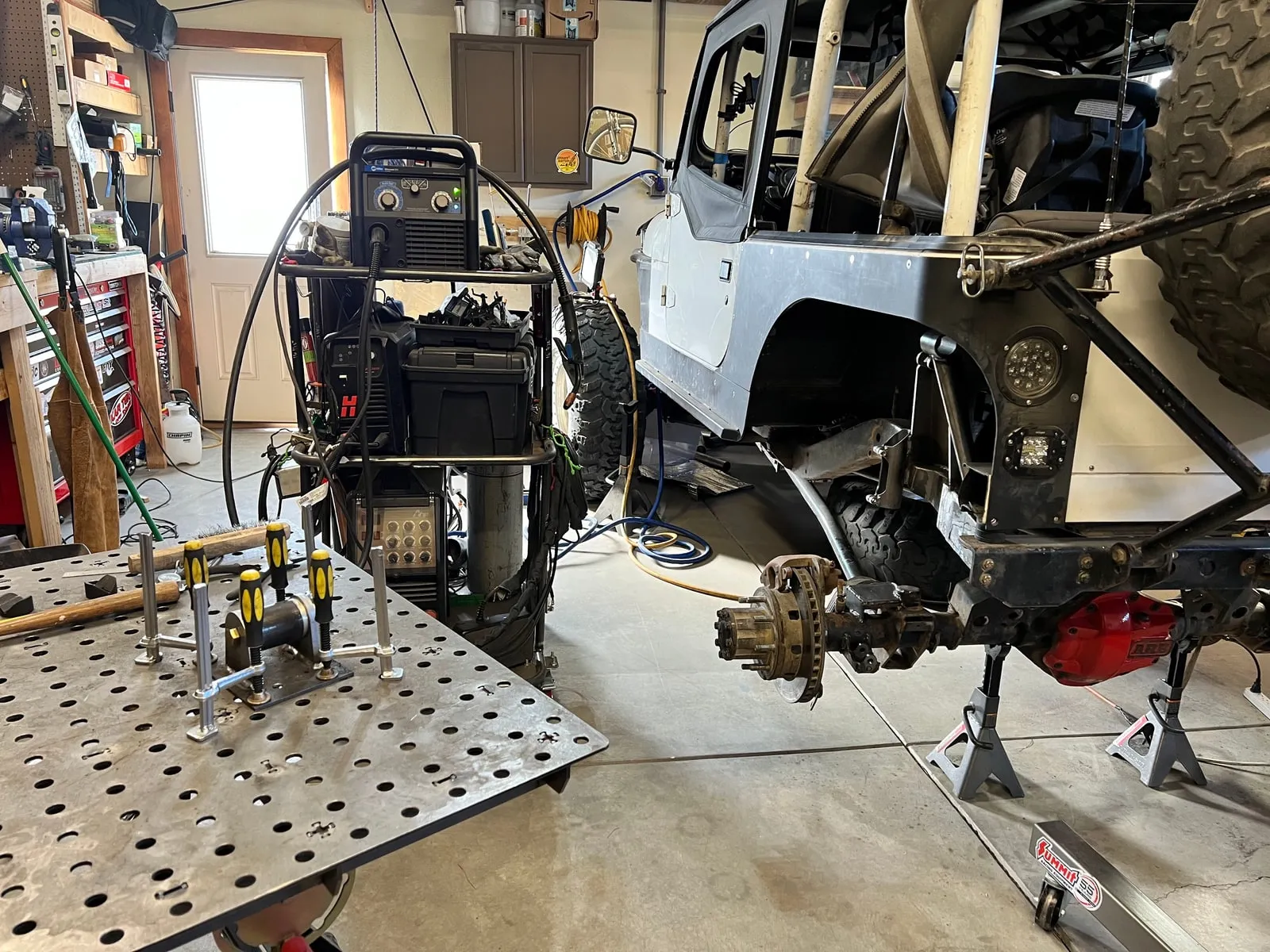 Alex Sulgrove's Jeep up on stands in his home shop, mid-fabrication with the rear shock mounts pulled and welding fixtures on the table.